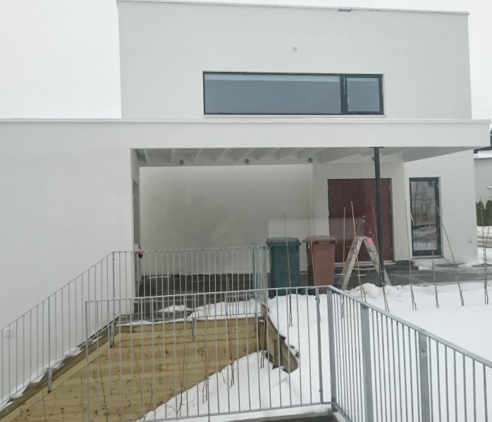 Nowoczesny, biały dom szkieletowy z płaskim dachem, widoczny fragment tarasu z metalową barierką i schodami. Na podjeździe stoją dwa kosze na śmieci i drabina.