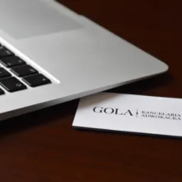 Wizytówka Kancelarii Adwokackiej GOLA leżąca obok srebrnego laptopa na ciemnym, drewnianym biurku.
