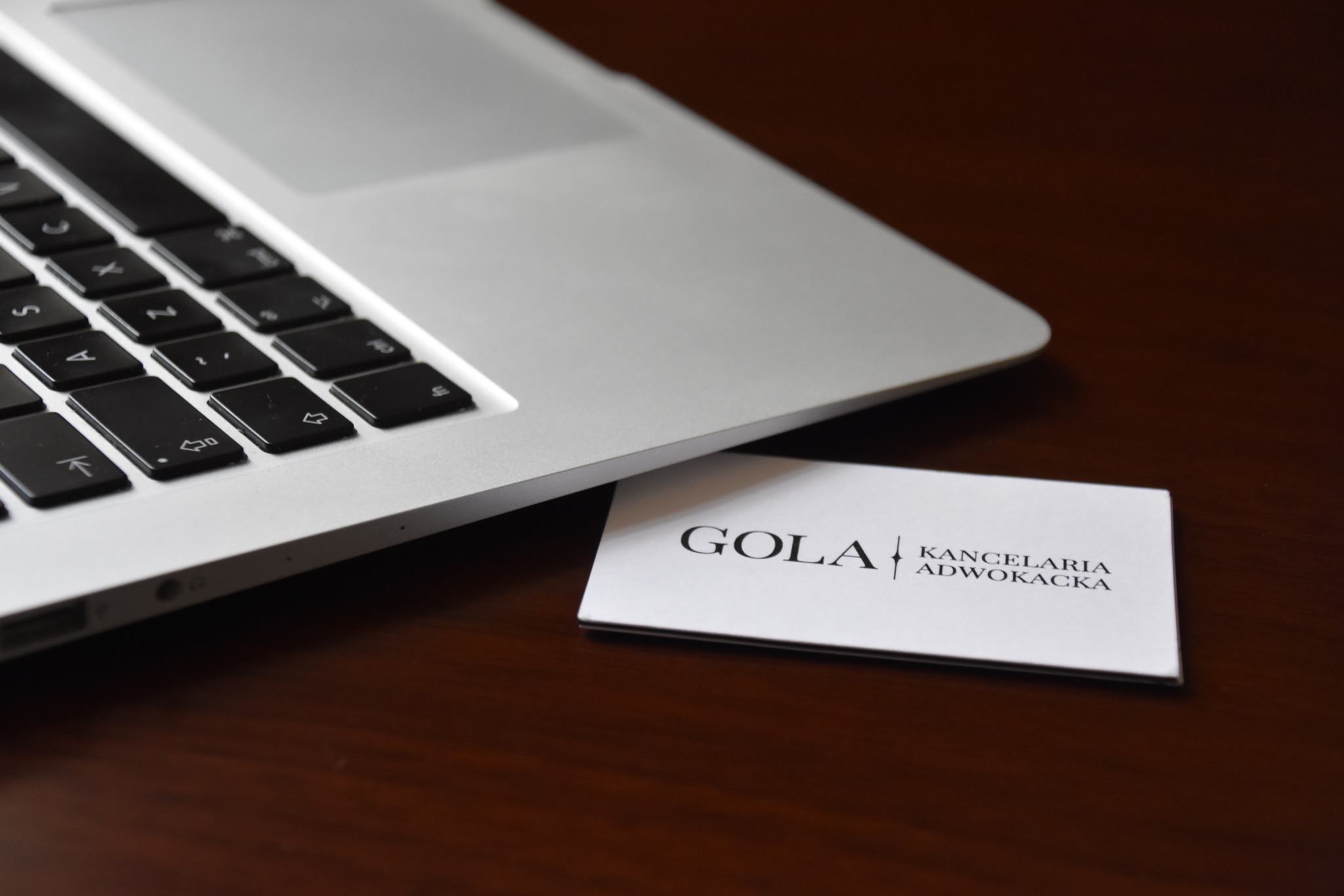 Wizytówka Kancelarii Adwokackiej GOLA leżąca obok srebrnego laptopa na ciemnym, drewnianym biurku.