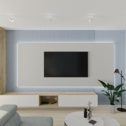 KT Studio Wnętrz Kamila Toruń - Nowoczesny salon z telewizorem na ścianie z podświetleniem LED, białe meble, drewniane akcenty i roślina doniczkowa. Minimalistyczny design.