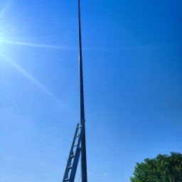 Electricity - Słup oświetleniowy z drabiną opartą o niego na tle błękitnego nieba i fragmentu krajobrazu z zielenią.