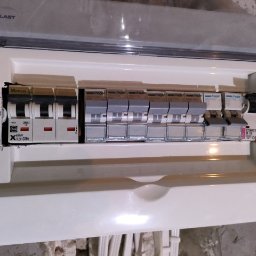 Electricity - Otwarta skrzynka elektryczna z zainstalowanymi bezpiecznikami, widoczne marki Hager i Moeller, częściowo zakryta plastikową osłoną Elektro-Plast.