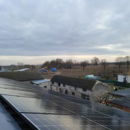 Electricity - Panele fotowoltaiczne na dachu budynku gospodarczego, w tle zabudowania wiejskie i pochmurne niebo.