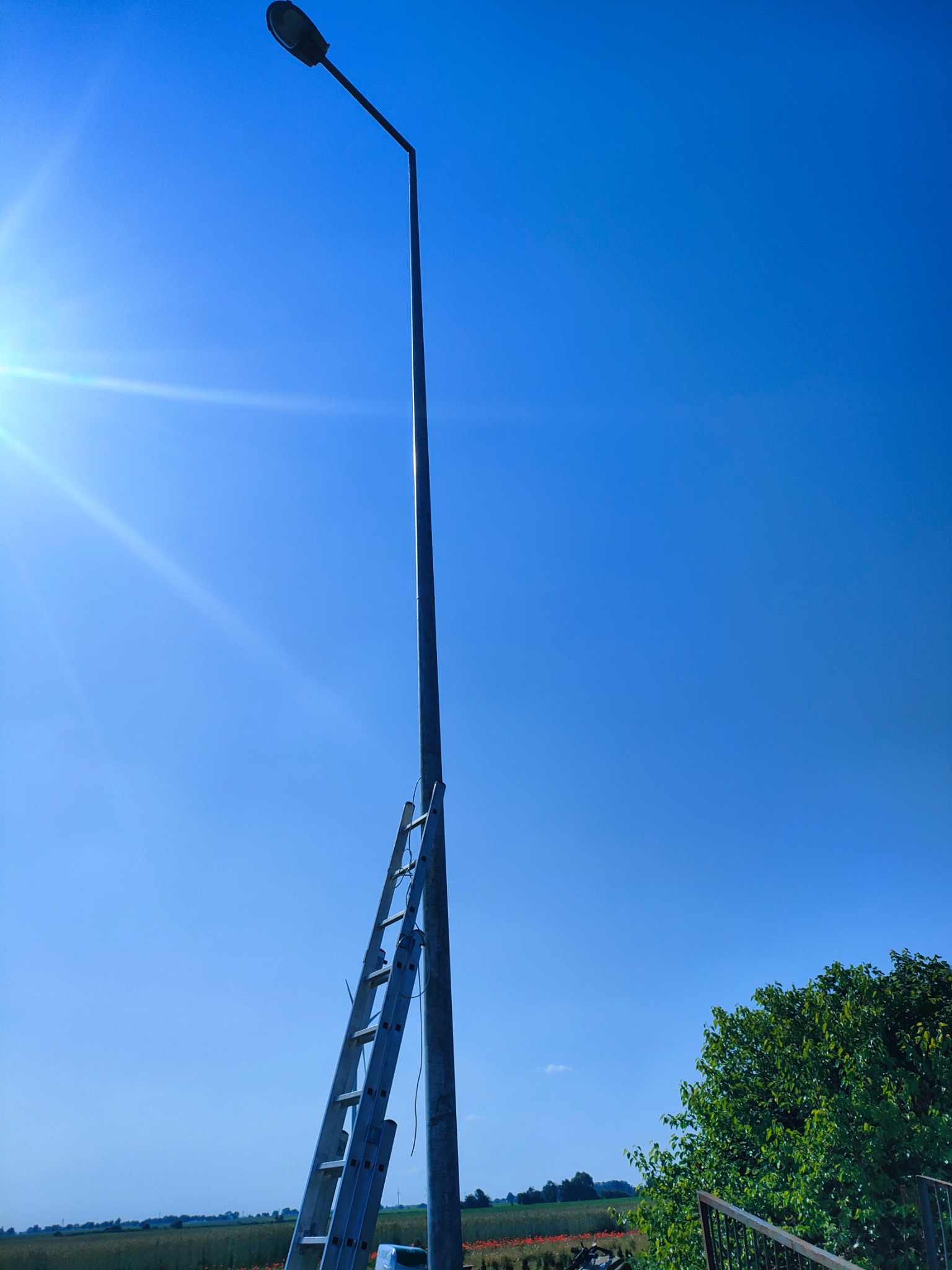 Słup oświetleniowy z drabiną opartą o niego na tle błękitnego nieba i fragmentu krajobrazu z zielenią.