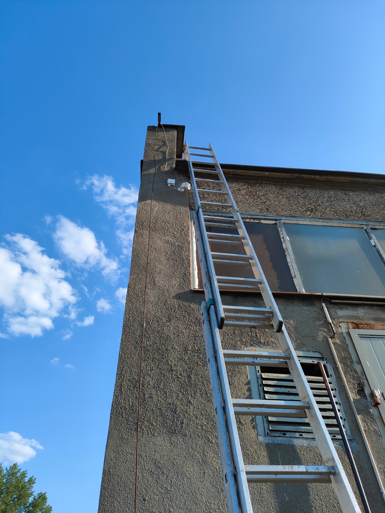 Aluminiowa drabina oparta o szorstką ścianę budynku z widocznymi oknami i kamerami monitoringu pod błękitnym niebem z chmurami.