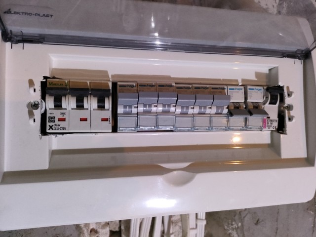 Otwarta skrzynka elektryczna z zainstalowanymi bezpiecznikami, widoczne marki Hager i Moeller, częściowo zakryta plastikową osłoną Elektro-Plast.