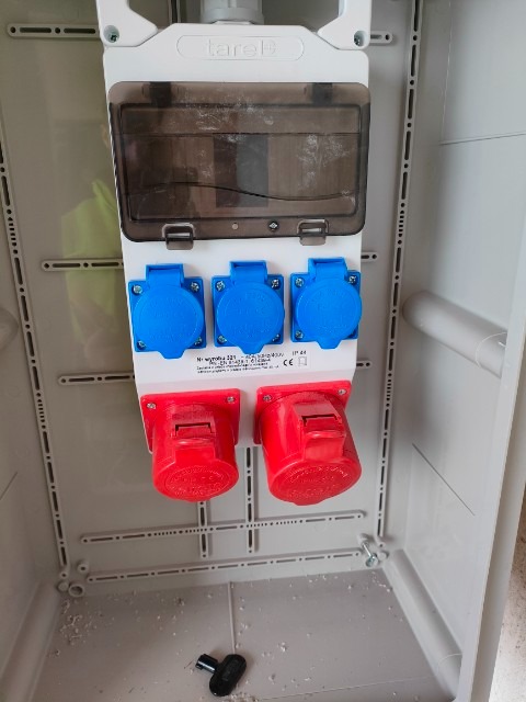 Skrzynka elektryczna z trzema niebieskimi i dwoma czerwonymi gniazdami, widoczna wewnątrz szarej obudowy, z kluczem leżącym na dnie.