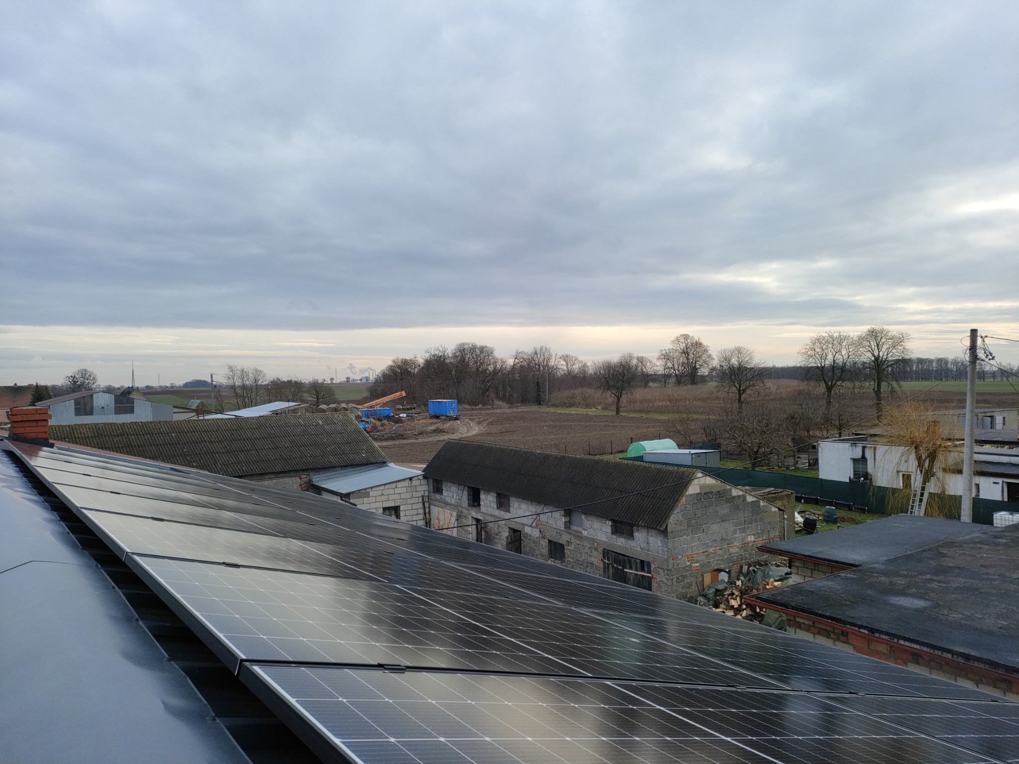 Panele fotowoltaiczne na dachu budynku gospodarczego, w tle zabudowania wiejskie i pochmurne niebo.