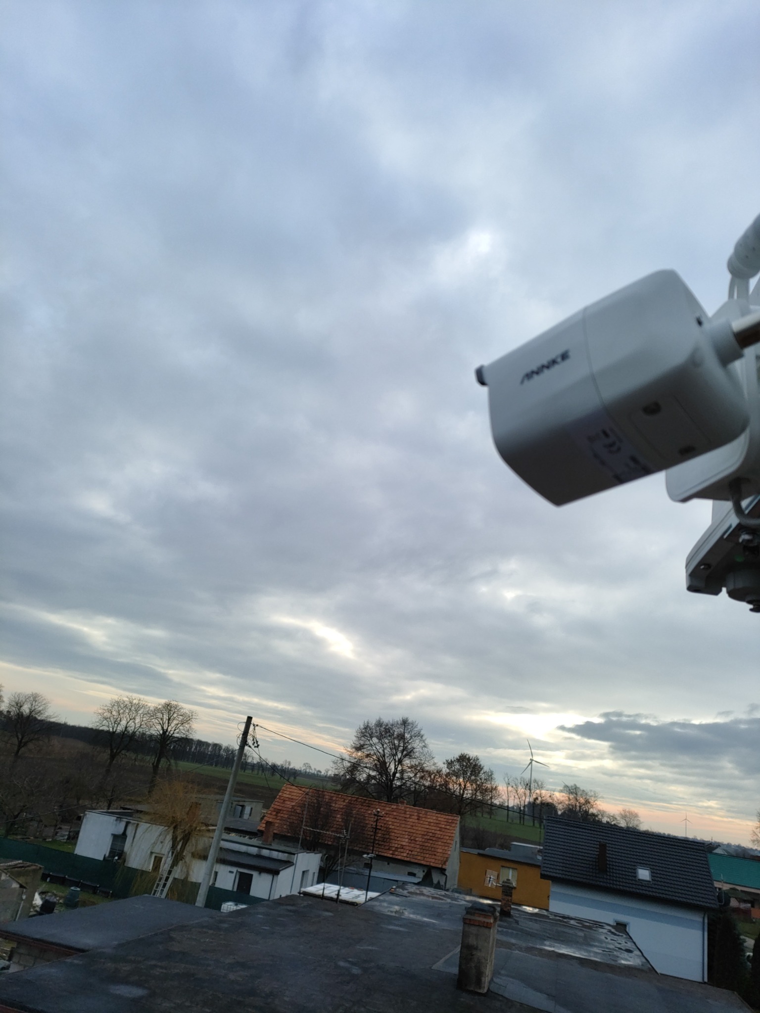 Biała kamera monitoringu marki ANKKE zamontowana na zewnątrz, z widokiem na wiejskie zabudowania i pochmurne niebo o zmierzchu.