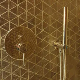 Błyszcząca, złota armatura prysznicowa zamontowana na ścianie wyłożonej trójkątnymi płytkami w odcieniach złota, tworzącymi geometryczny wzór.