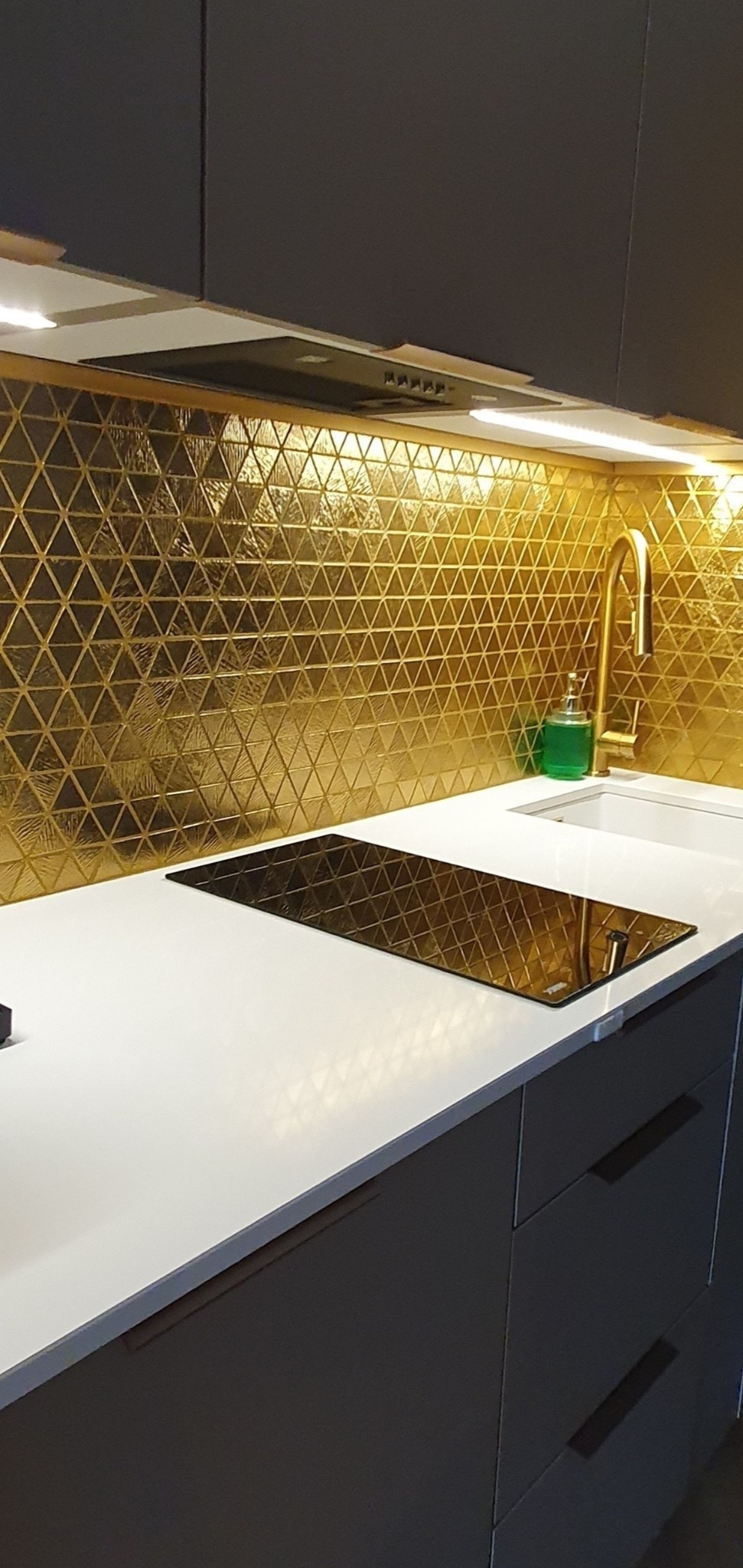 Nowoczesna kuchnia z błyszczącą, złotą mozaiką trójkątną na ścianie nad blatem, ciemnoszare szafki z minimalistycznymi uchwytami i biały blat z płytą indukcyjną.