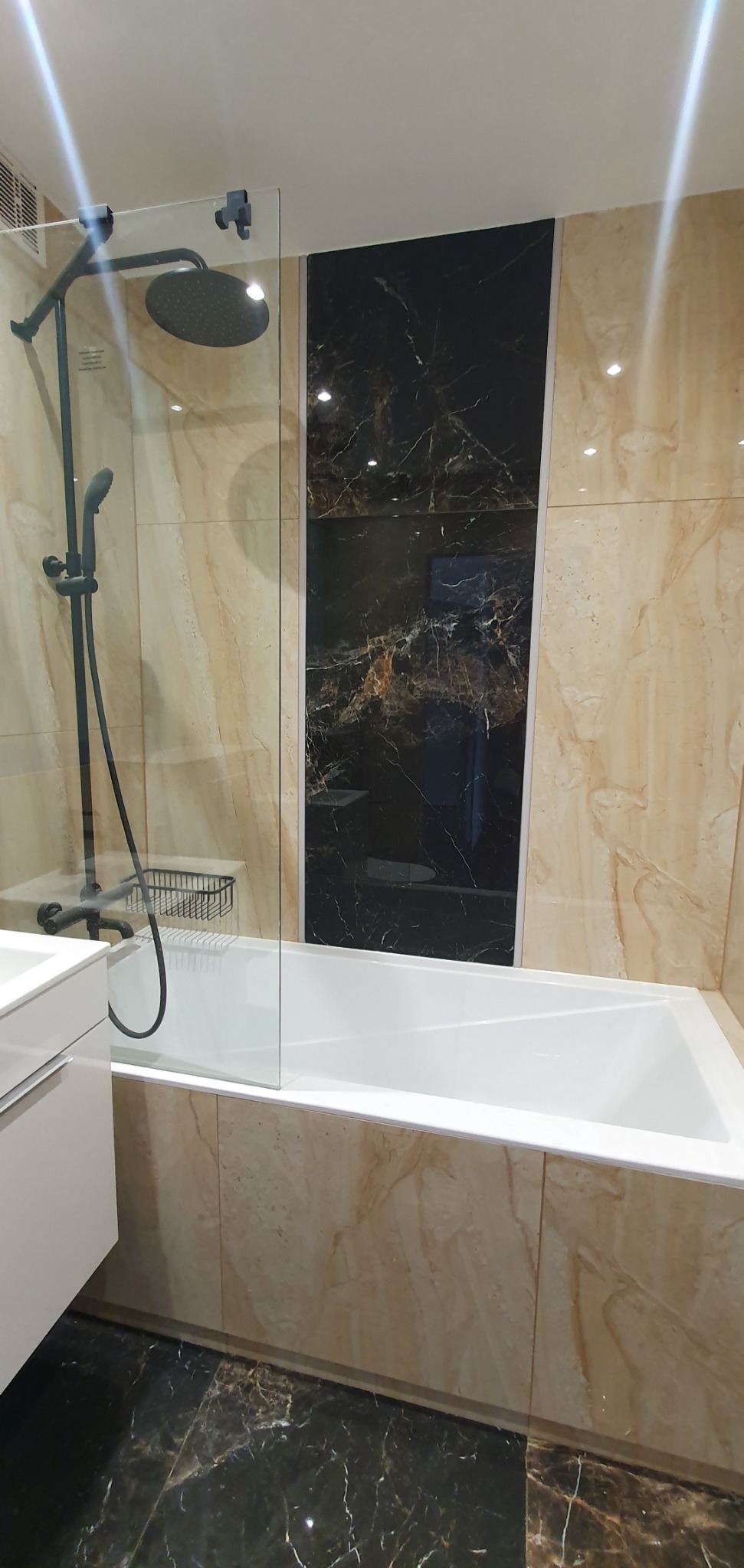 Nowoczesna łazienka z wanną obudowaną beżowymi płytkami imitującymi marmur, czarny marmurowy panel na ścianie, szklany panel prysznicowy z czarną deszczownicą i baterią.