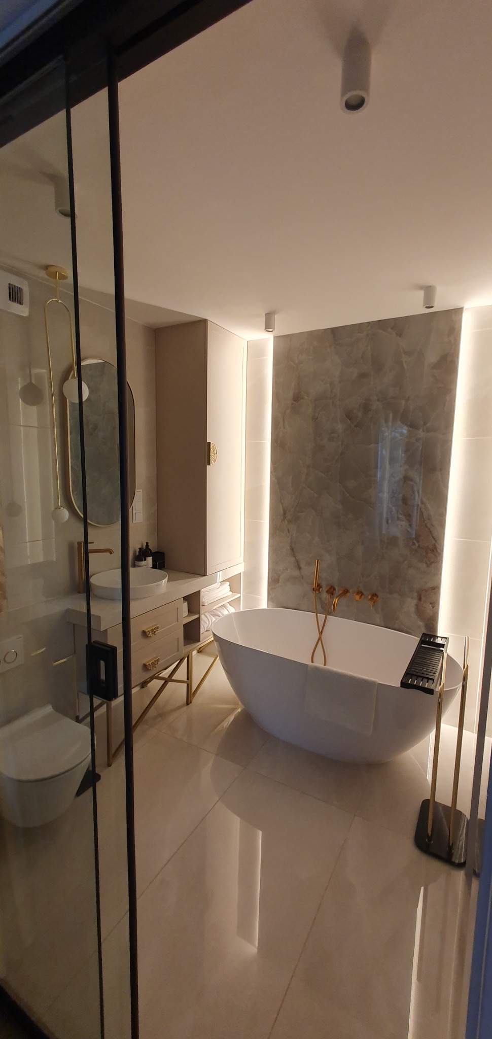 Elegancka łazienka z wolnostojącą wanną, marmurową ścianą z podświetleniem LED, umywalką na szafce z złotymi nogami i armaturą oraz szklaną kabiną prysznicową.