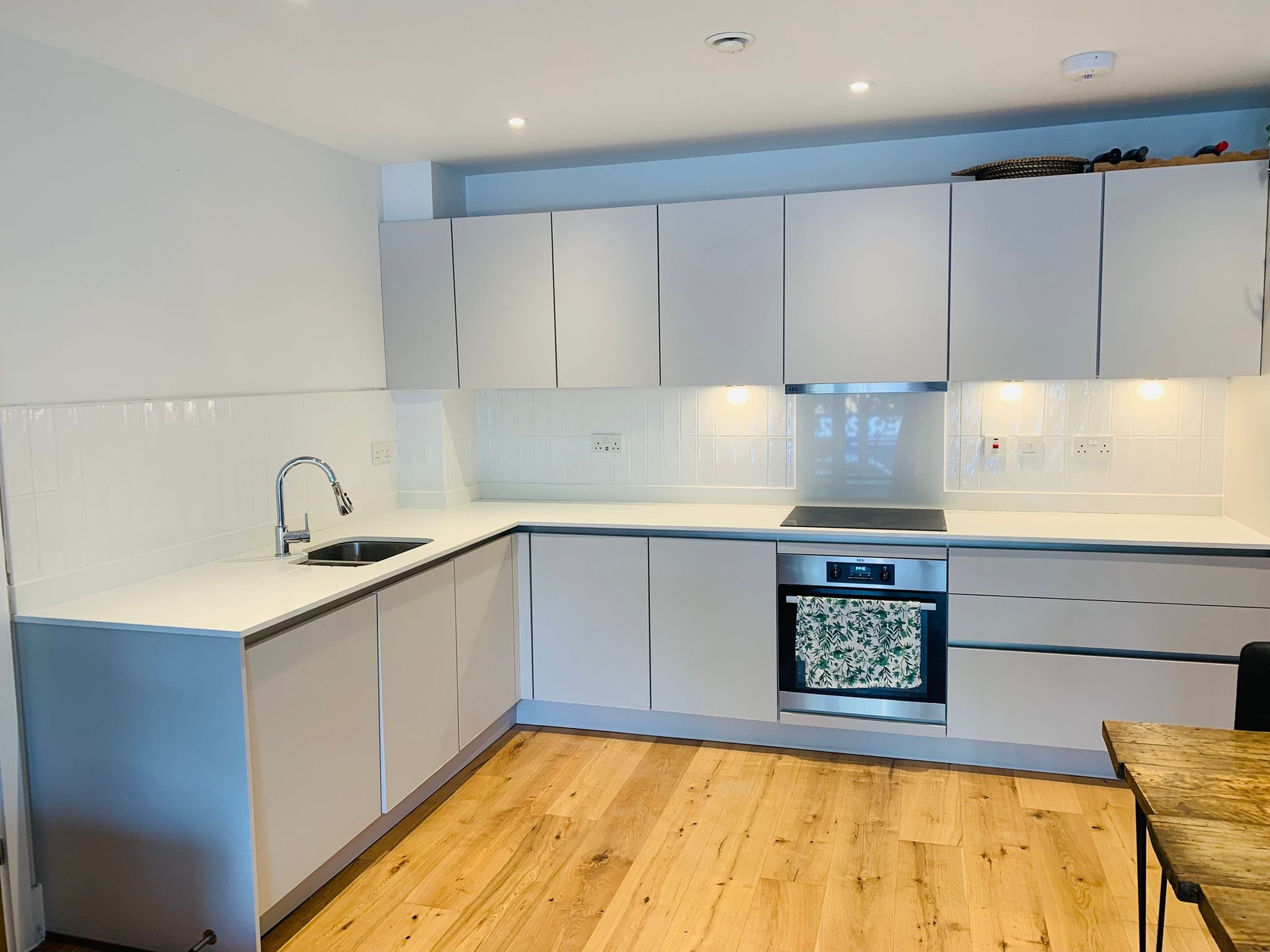 Jasna, nowoczesna kuchnia z drewnianą podłogą; widoczne białe szafki, blat, zlewozmywak, płyta indukcyjna i piekarnik z ręcznikiem w kwiaty na drzwiach.