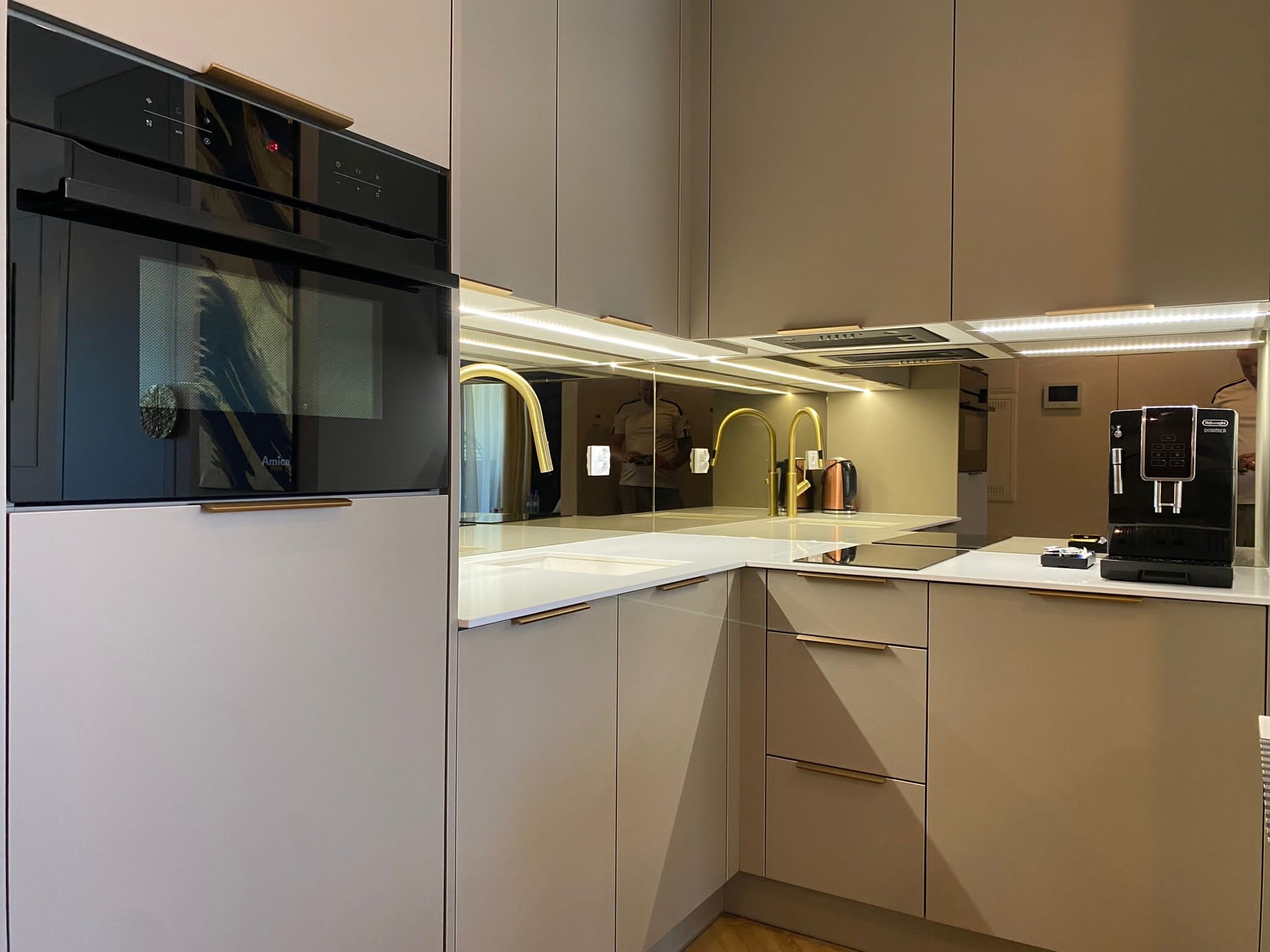 Nowoczesna kuchnia z beżowymi szafkami, złotymi uchwytami, lustrzanym panelem nad blatem i wbudowanym piekarnikiem marki Amica oraz ekspresem do kawy DeLonghi.