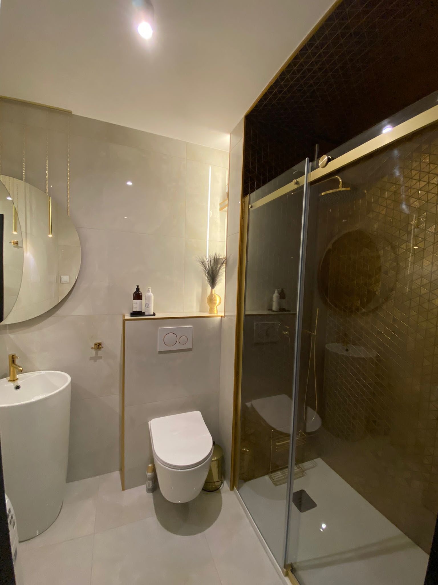 Nowoczesna łazienka z kabiną prysznicową z geometrycznym wzorem na ścianie, wiszącą umywalką i toaletą, podkreślona złotymi detalami.