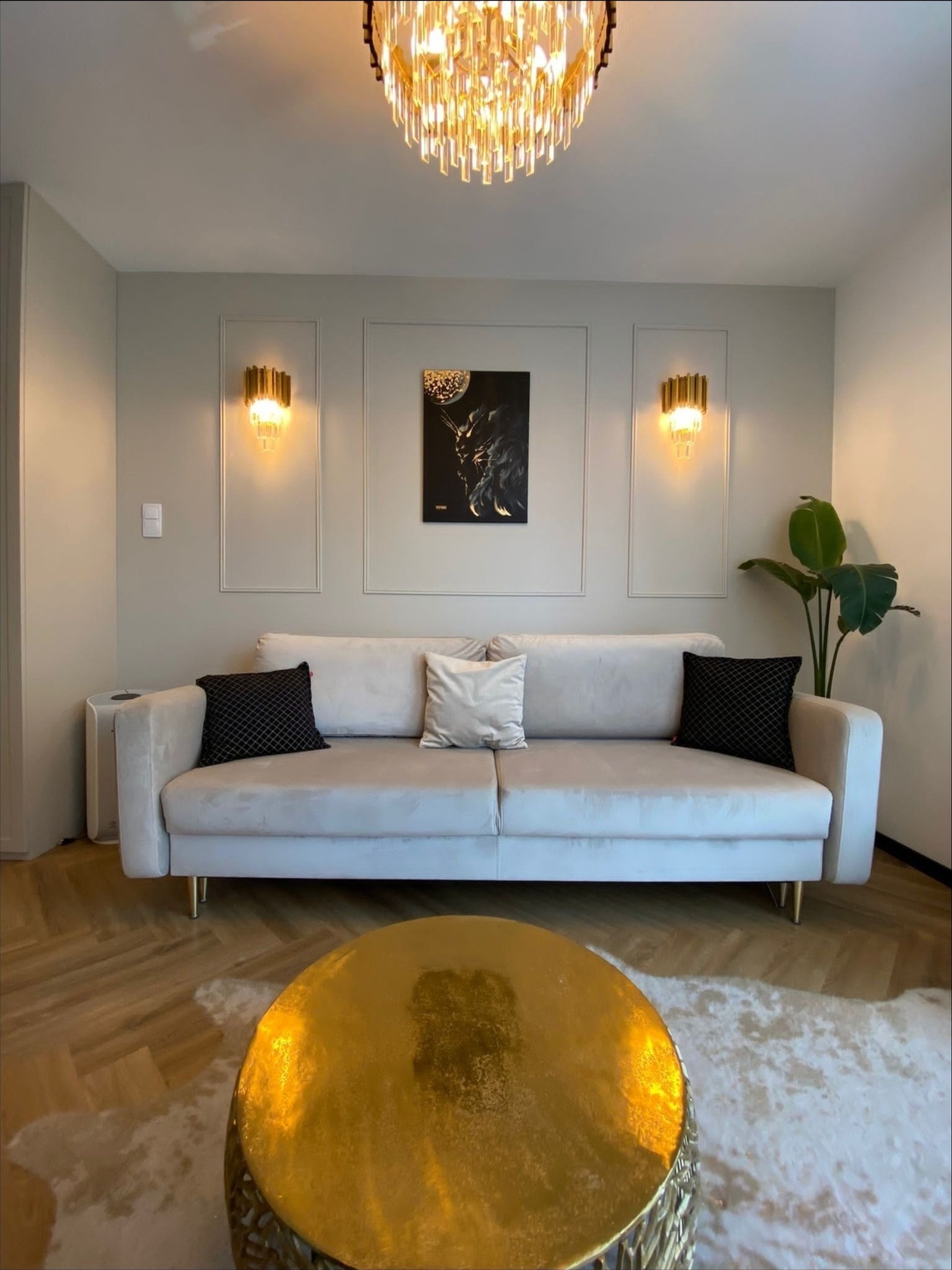 Elegancki salon z beżową sofą, złotym stolikiem kawowym, parkietem w jodełkę i designerskim oświetleniem ściennym i sufitowym.