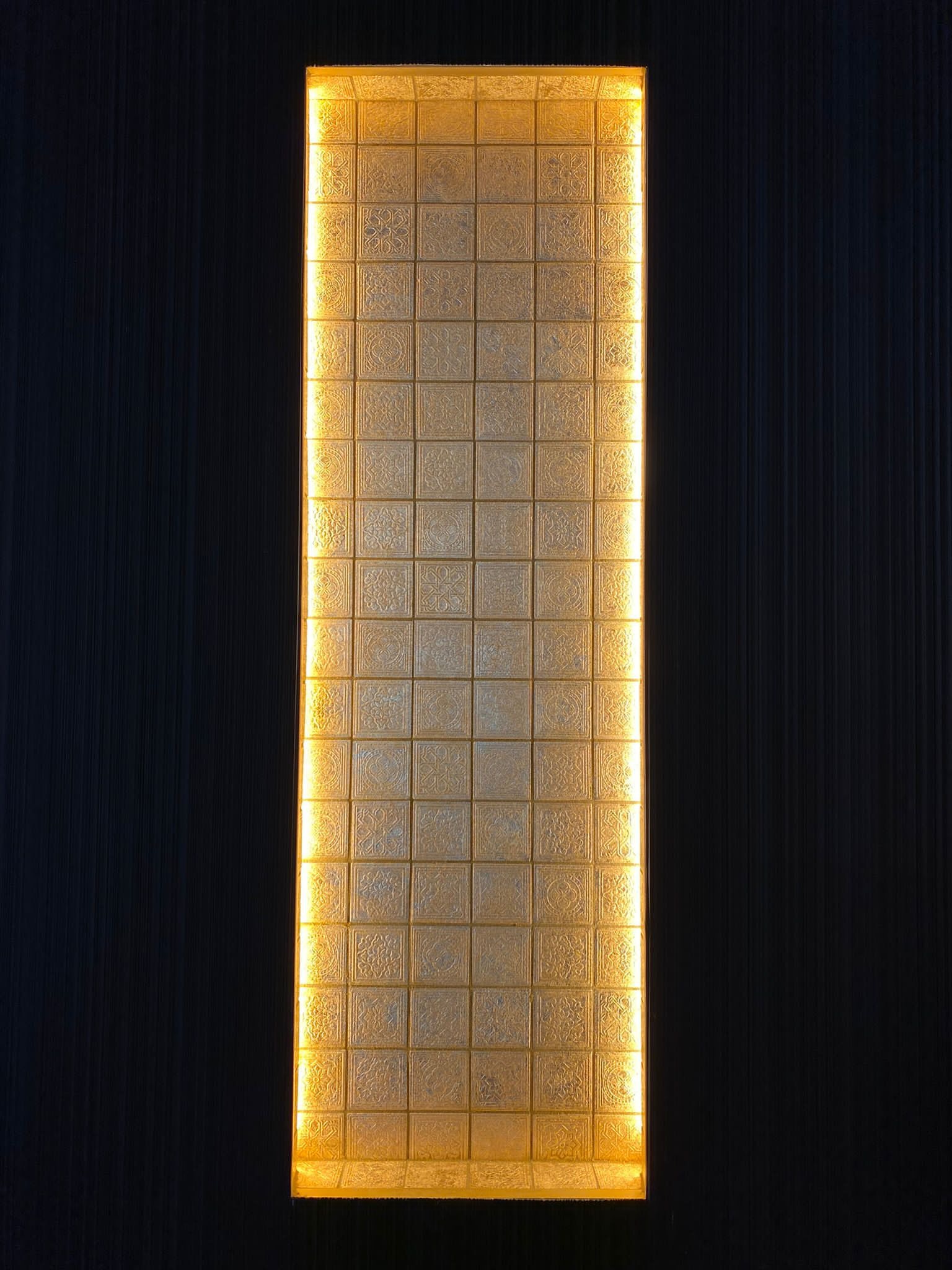Podświetlona dekoracyjna ściana z kwadratowych paneli o złocistym odcieniu z ornamentami, przylegająca do ciemnej, pionowo żłobkowanej ściany.