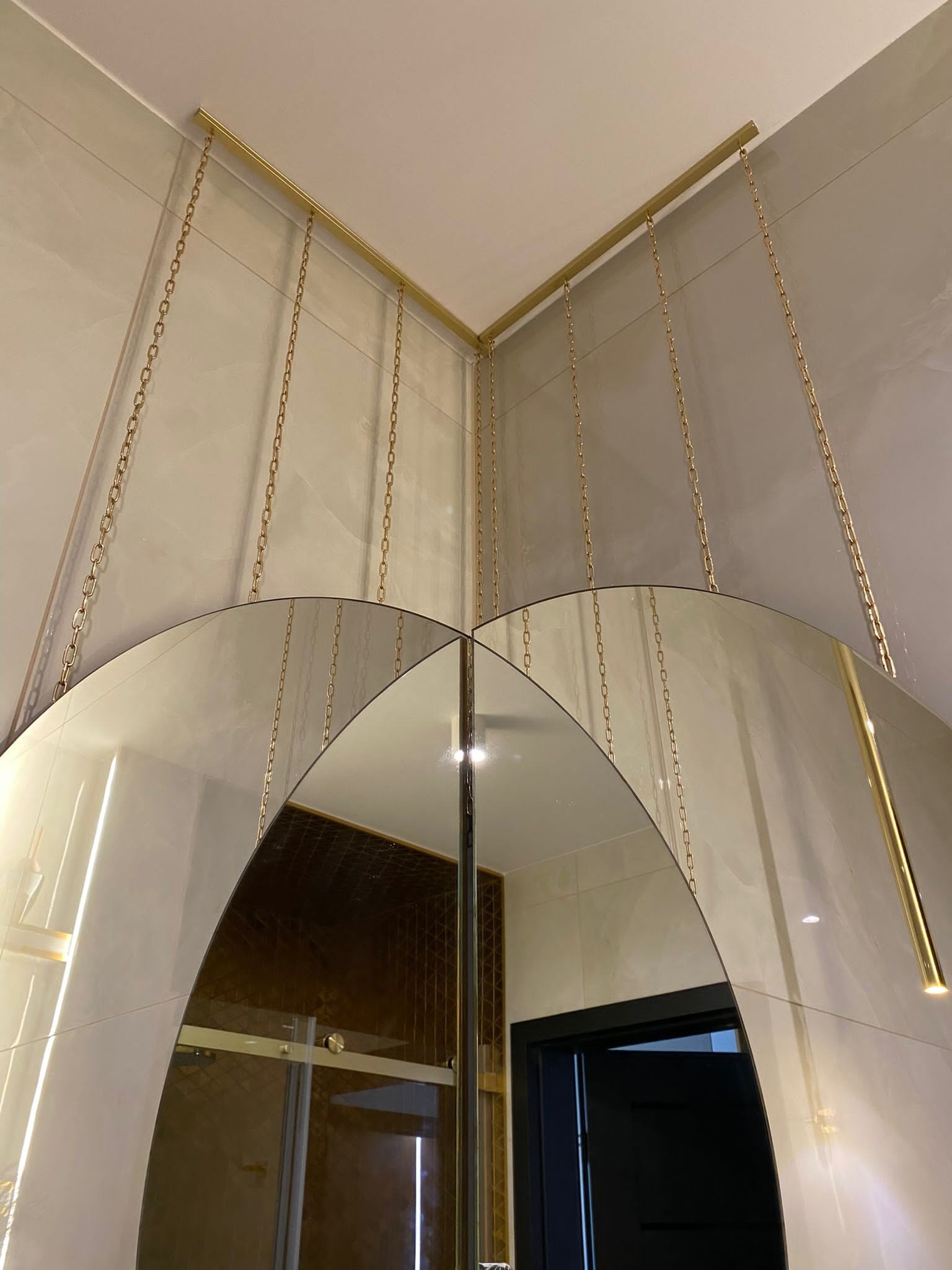 Nowoczesna łazienka z dwoma półokrągłymi lustrami zawieszonymi na złotych łańcuchach, podwieszonych do złotej ramy, z widoczną kabiną prysznicową w tle.