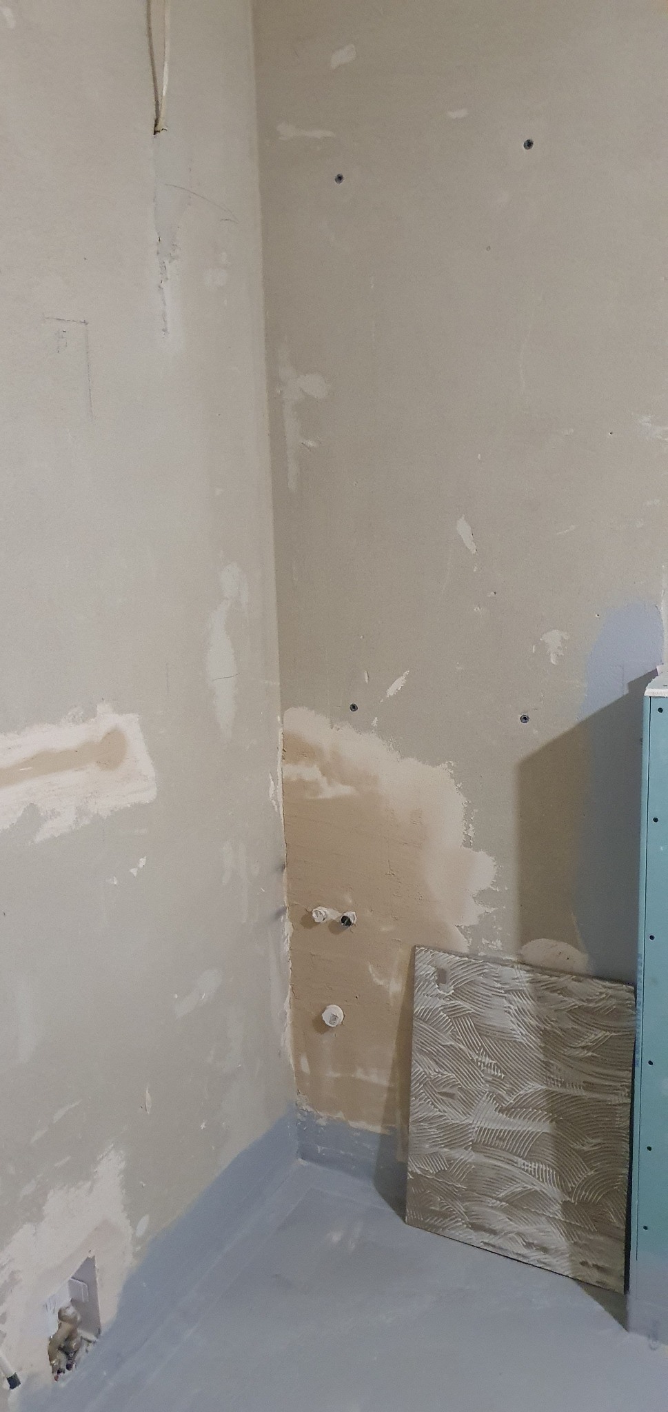 Przygotowanie ścian do położenia płytek w łazience: zagruntowana podłoga, fragment ściany z instalacją hydrauliczną, płyta gipsowo-kartonowa i płytka ceramiczna oparta o ścianę.