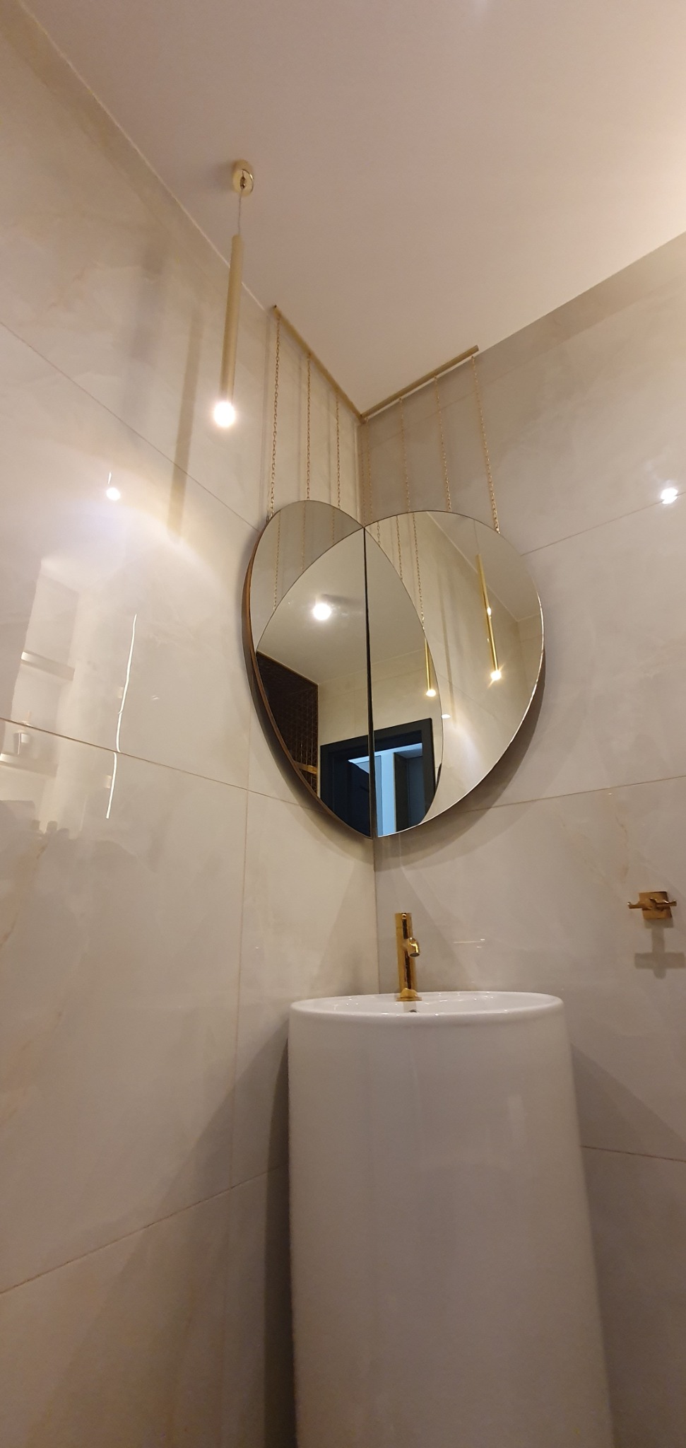 Nowoczesna łazienka z umywalką na postumencie, złotą armaturą i lustrem w kształcie serca zawieszonym na łańcuszkach, z widocznym oświetleniem punktowym.