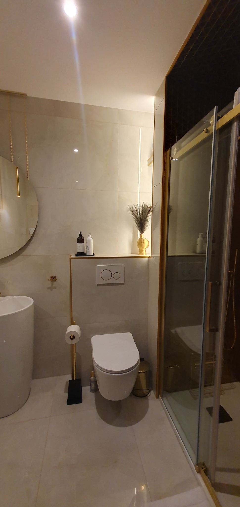 Elegancka łazienka z beżowymi płytkami, złotymi akcentami i kabiną prysznicową typu walk-in, toaleta wisząca, okrągłe lustro z podświetleniem i dekoracyjny wazon ze strusimi piórami.