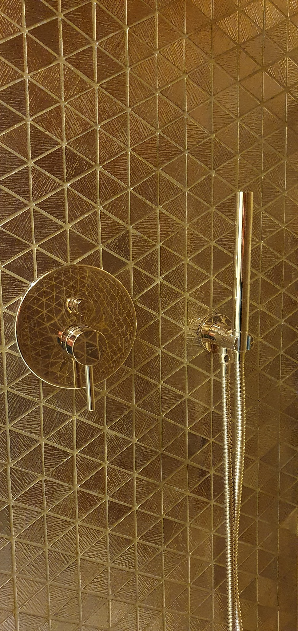 Błyszcząca, złota armatura prysznicowa zamontowana na ścianie wyłożonej trójkątnymi płytkami w odcieniach złota, tworzącymi geometryczny wzór.