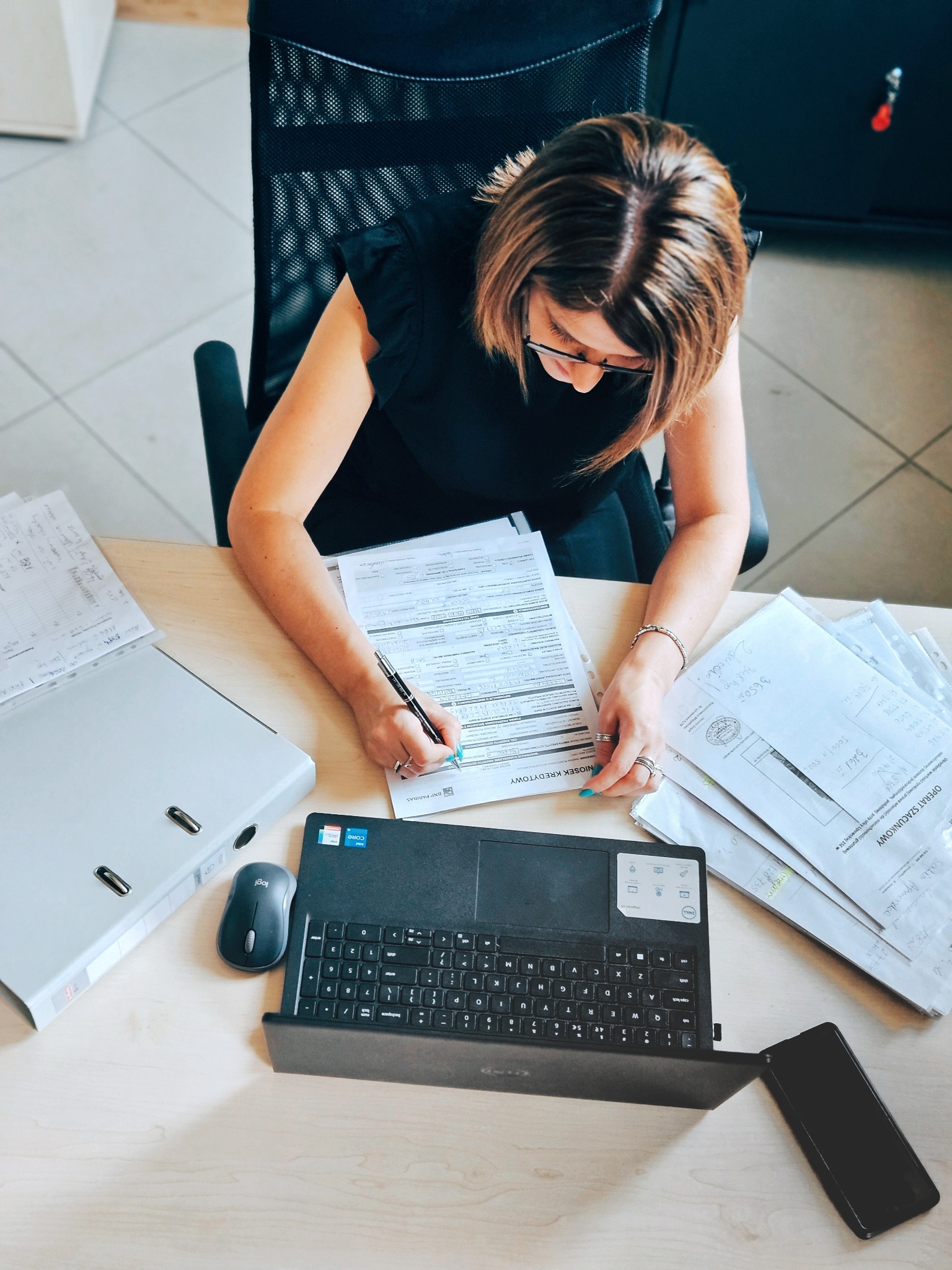 Kobieta w okularach wypełnia wniosek na drewnianym biurku, obok laptop, myszka, telefon i teczka z dokumentami.