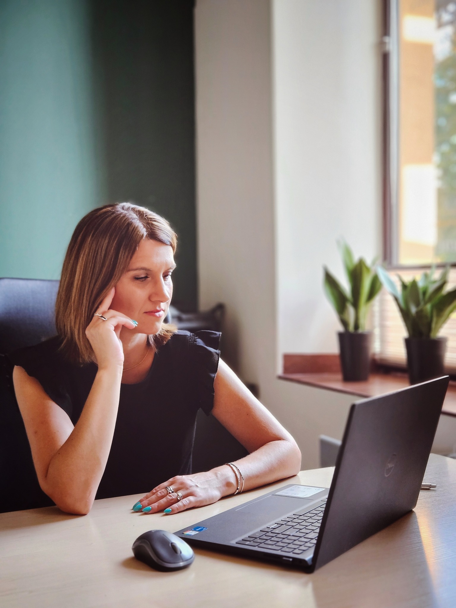 Kobieta w czarnej bluzce siedzi przy biurku z laptopem Dell, zamyślona z dłonią podpartą o policzek, widoczne rośliny w doniczkach na parapecie w tle.