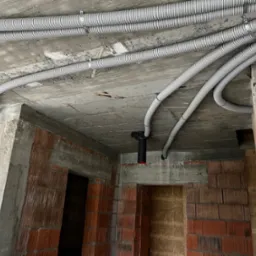 Szare, karbowane rury instalacyjne przymocowane do surowego, betonowego sufitu w budynku w stanie deweloperskim z widocznymi ścianami z czerwonej cegły.