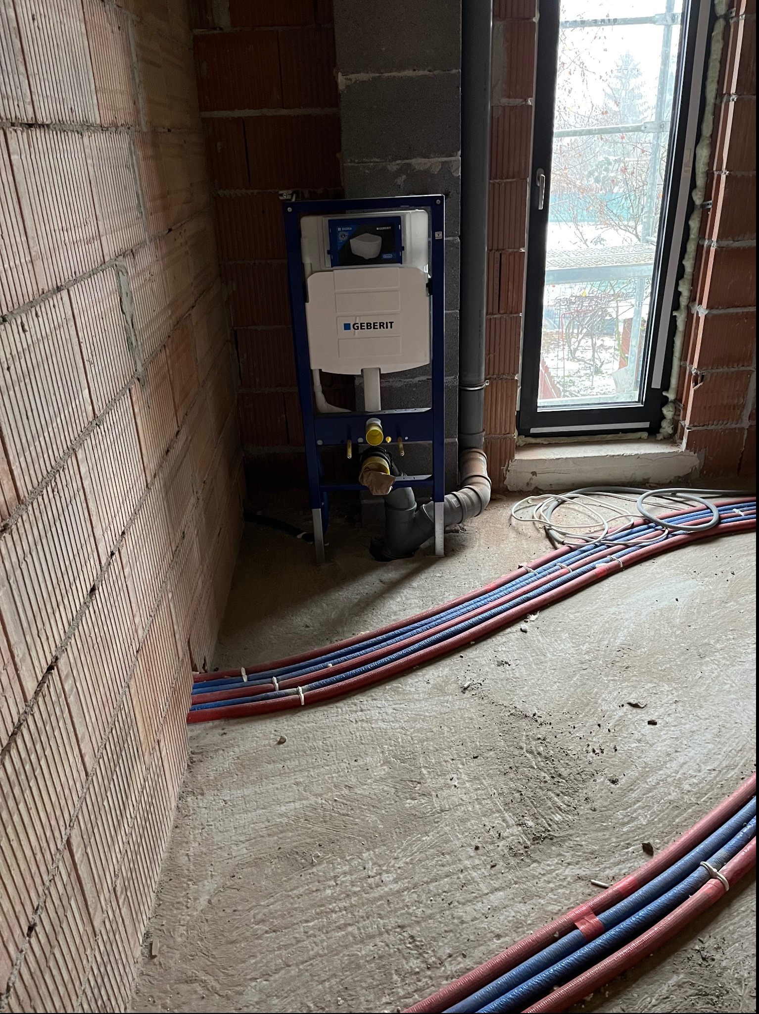 Stelaż podtynkowy Geberit zamontowany przy ceglanej ścianie w surowym stanie, widoczne rury instalacji wodnej i kanalizacyjnej, okno w tle.