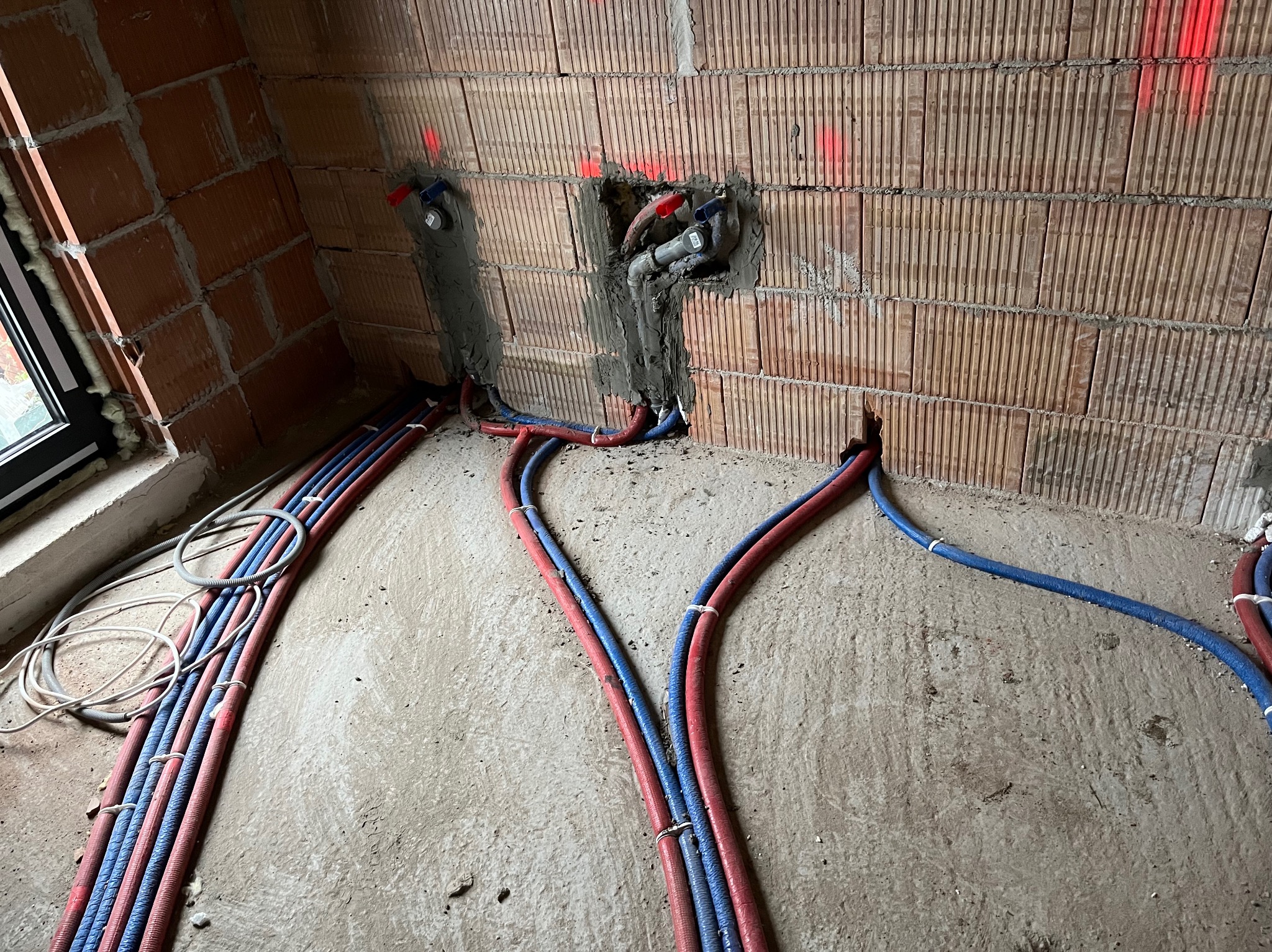 Instalacja wodna i grzewcza w trakcie montażu w surowym budynku, widoczne rury w izolacji czerwonej i niebieskiej, bruzdy w ścianach z cegły oraz wystające przyłącza.