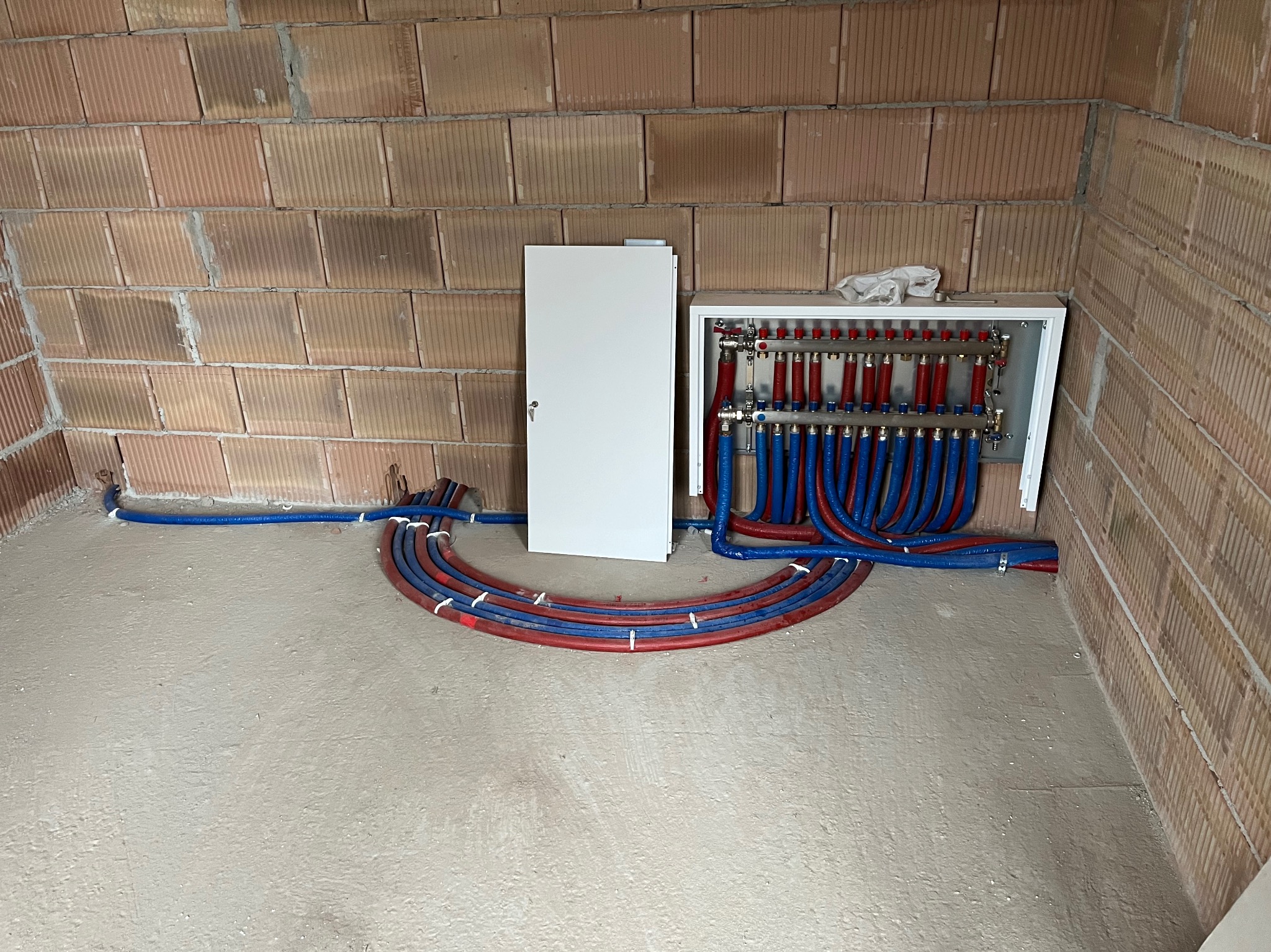 Rozdzielacz ogrzewania podłogowego z rurami w kolorze czerwonym i niebieskim, ułożonymi na świeżym betonie w surowym wnętrzu z ceglanymi ścianami.