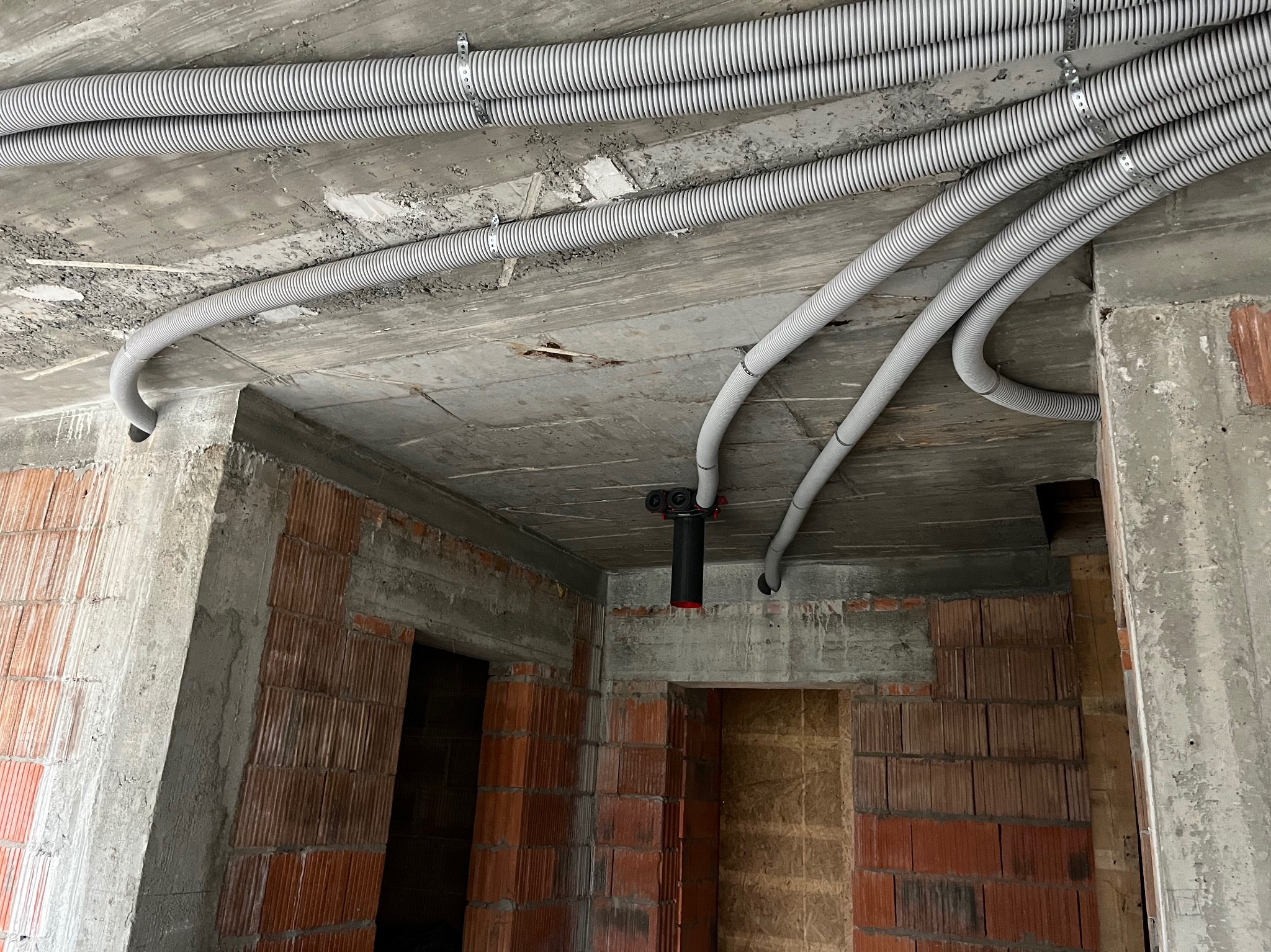 Szare, karbowane rury instalacyjne przymocowane do surowego, betonowego sufitu w budynku w stanie deweloperskim z widocznymi ścianami z czerwonej cegły.
