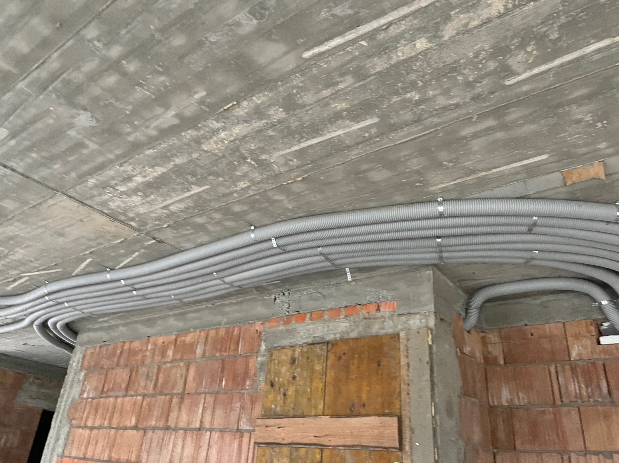 Szare rury instalacyjne przymocowane do sufitu w stanie surowym w Łukowej, widoczne cegły i beton, instalacja elektryczna w trakcie wykonywania.