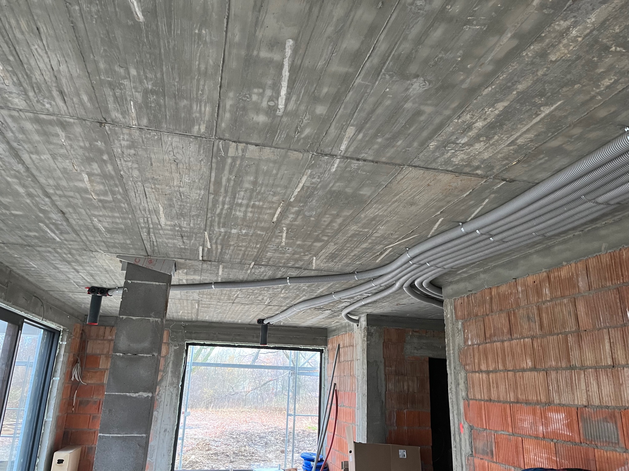 Szare, karbowane rury instalacyjne prowadzone pod betonowym stropem w surowym stanie, widoczne ceglane ściany i okna na zewnątrz.