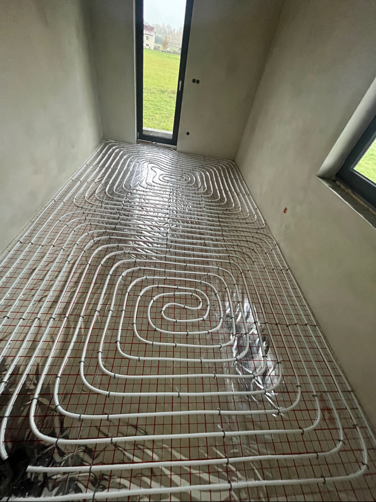 Białe rury ogrzewania podłogowego ułożone spiralnie na czerwonej siatce w surowym pomieszczeniu z oknem na zewnątrz.