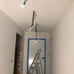 Usługi ogólnobudowlane MJ Marian Jakim - Korytarz w trakcie prac wykończeniowych: świeżo wyszpachlowane ściany, zabezpieczone taśmą drzwi, widoczne przewody elektryczne i drabina aluminiowa.