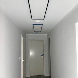 Usługi ogólnobudowlane MJ Marian Jakim - Korytarz w trakcie malowania na szaro, z widocznymi panelami sufitowymi z czarną ramką, zabezpieczonymi taśmą malarską, oraz przewodami na podłodze.