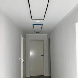 Korytarz w trakcie malowania na szaro, z widocznymi panelami sufitowymi z czarną ramką, zabezpieczonymi taśmą malarską, oraz przewodami na podłodze.