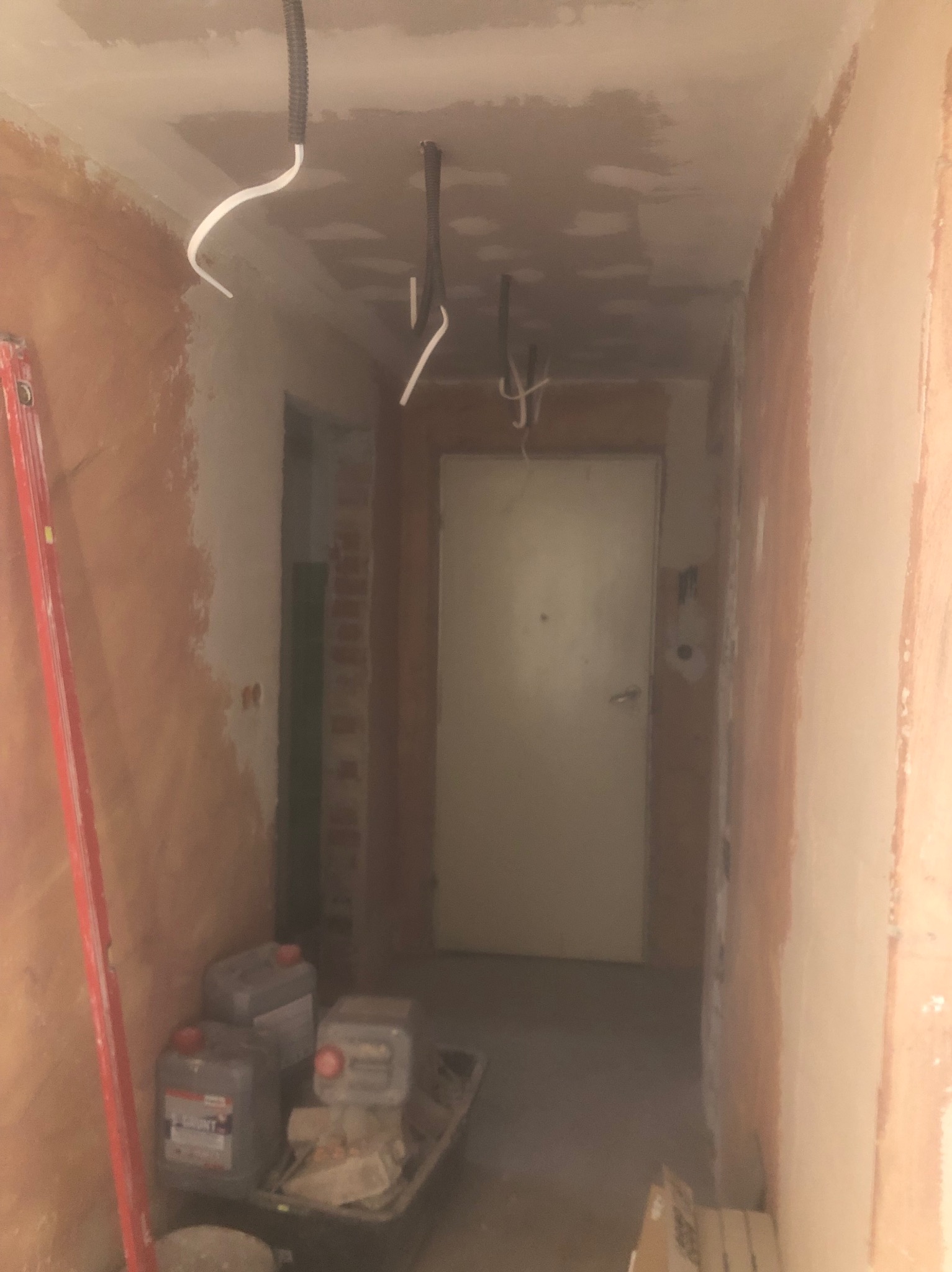 Korytarz w trakcie prac przygotowawczych do malowania, ściany częściowo pokryte gruntem, widoczne kable wystające z sufitu, drabina i pojemniki z farbą.