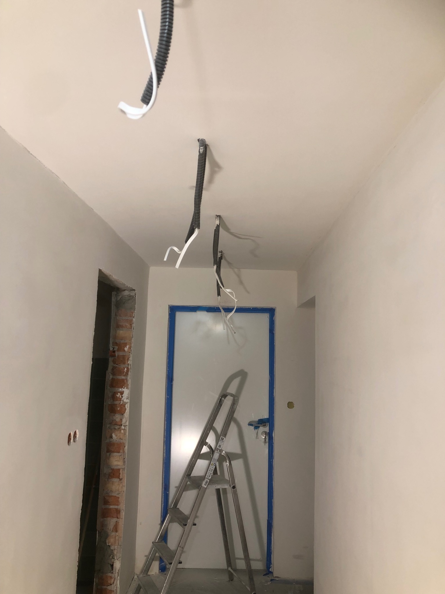 Korytarz w trakcie prac wykończeniowych: świeżo wyszpachlowane ściany, zabezpieczone taśmą drzwi, widoczne przewody elektryczne i drabina aluminiowa.