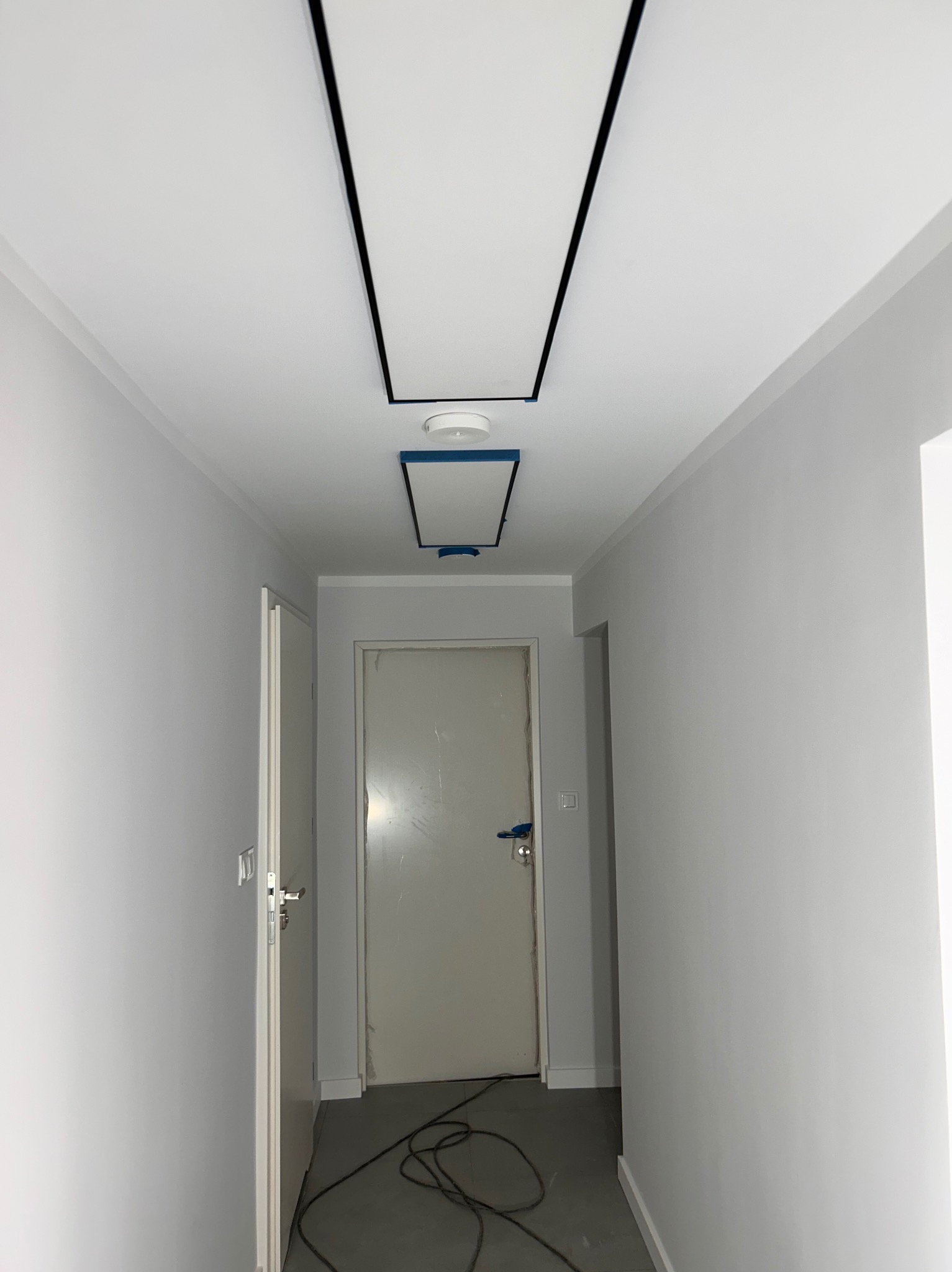 Korytarz w trakcie malowania na szaro, z widocznymi panelami sufitowymi z czarną ramką, zabezpieczonymi taśmą malarską, oraz przewodami na podłodze.