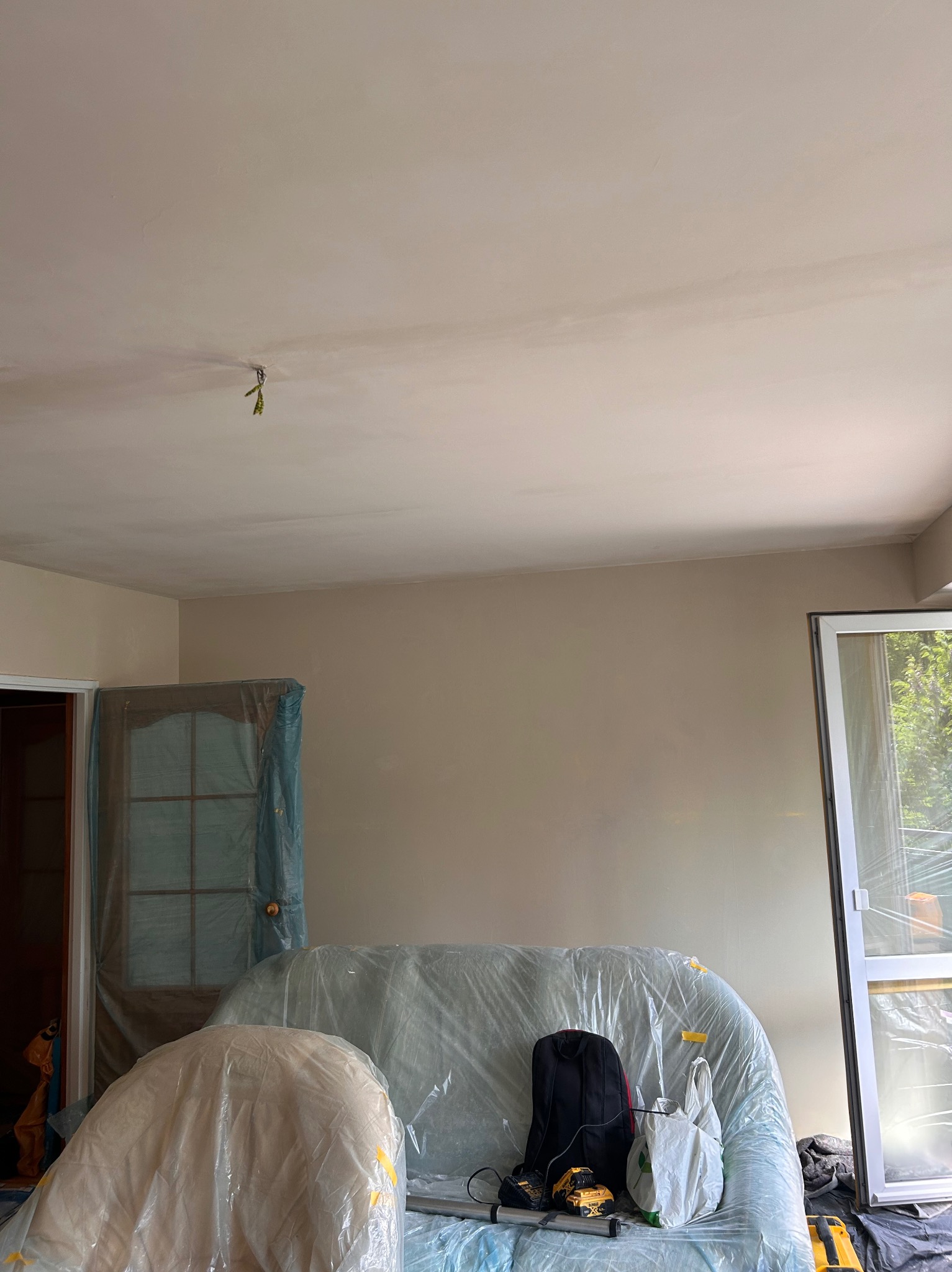 Pokój w trakcie przygotowania do malowania: meble zakryte folią, świeżo zagipsowana ściana, widoczne narzędzia i torba.