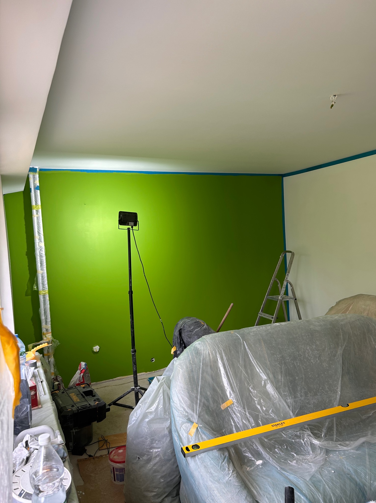 Pokój w trakcie malowania na jaskrawy zielony kolor, zabezpieczone listwy przypodłogowe taśmą malarską, widoczny reflektor na statywie, drabina, meble przykryte folią ochronną i poziomica Stanley.