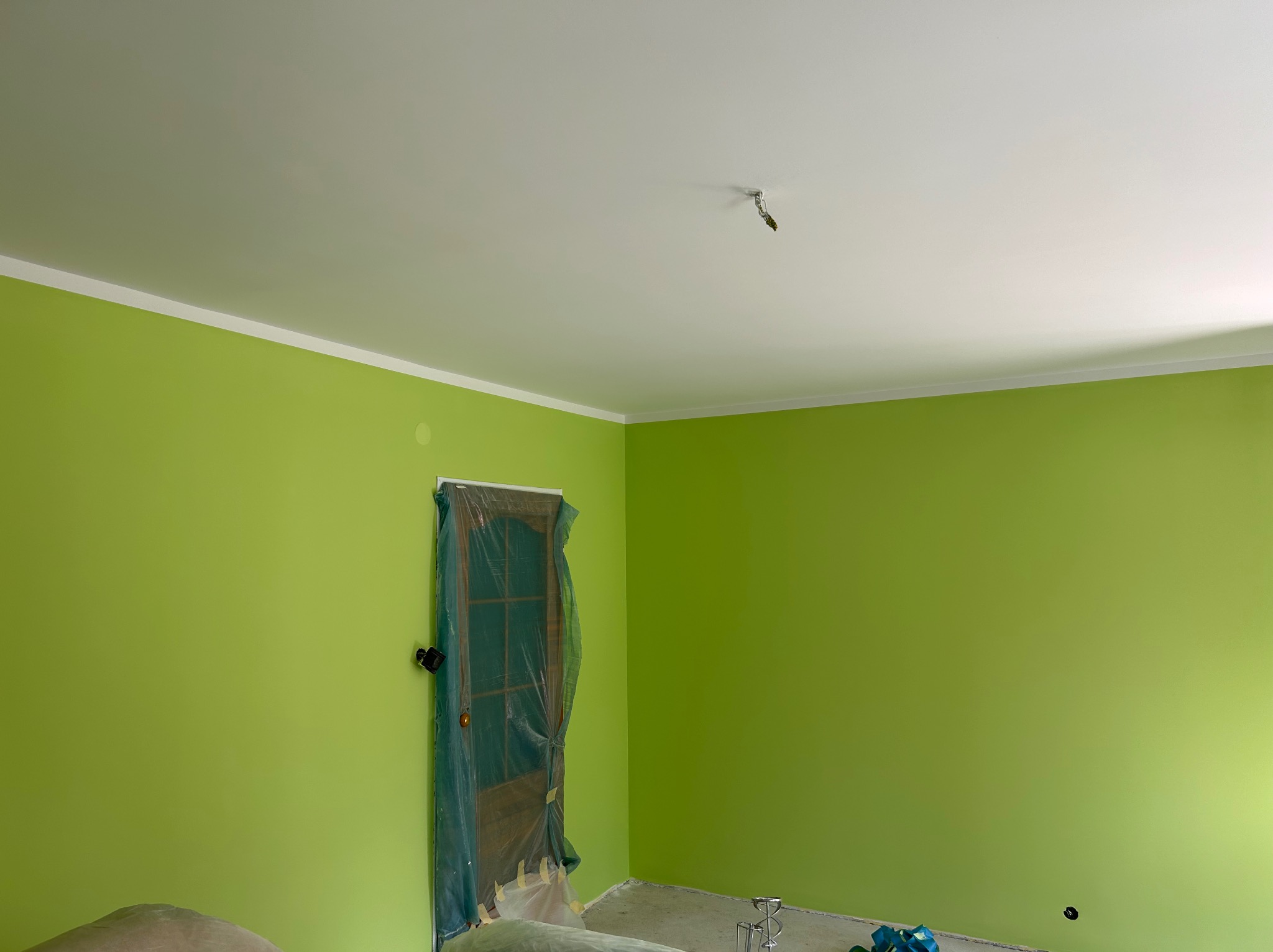 Świeżo pomalowane na zielono ściany pokoju z zabezpieczonymi drzwiami folią malarską; widoczny biały sufit z listwą przypodłogową i wystającym kablem od lampy.