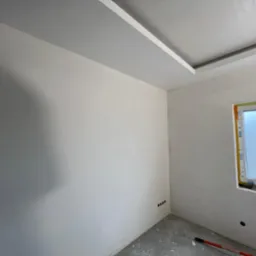 Gładkie, białe ściany po malowaniu w Koszalinie. Widoczne gniazdka elektryczne i okno z ramą. Na podłodze ślady farby i wałek malarski.