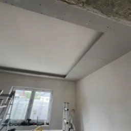 Montaż sufitu podwieszanego z płyt gipsowo-kartonowych w pomieszczeniu z oknem. Widoczne rusztowania i narzędzia. Prace wykończeniowe wnętrz w trakcie realizacji.