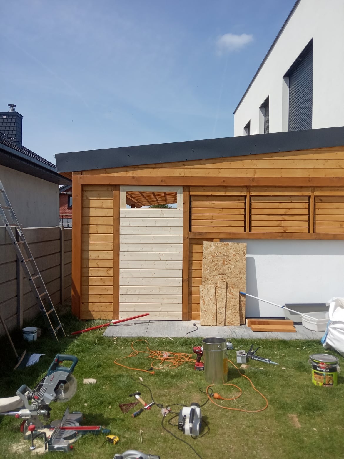 Drewniana konstrukcja z płaskim dachem w trakcie budowy, widoczne narzędzia, drabina i materiały budowlane na trawniku, w tle nowoczesny dom.