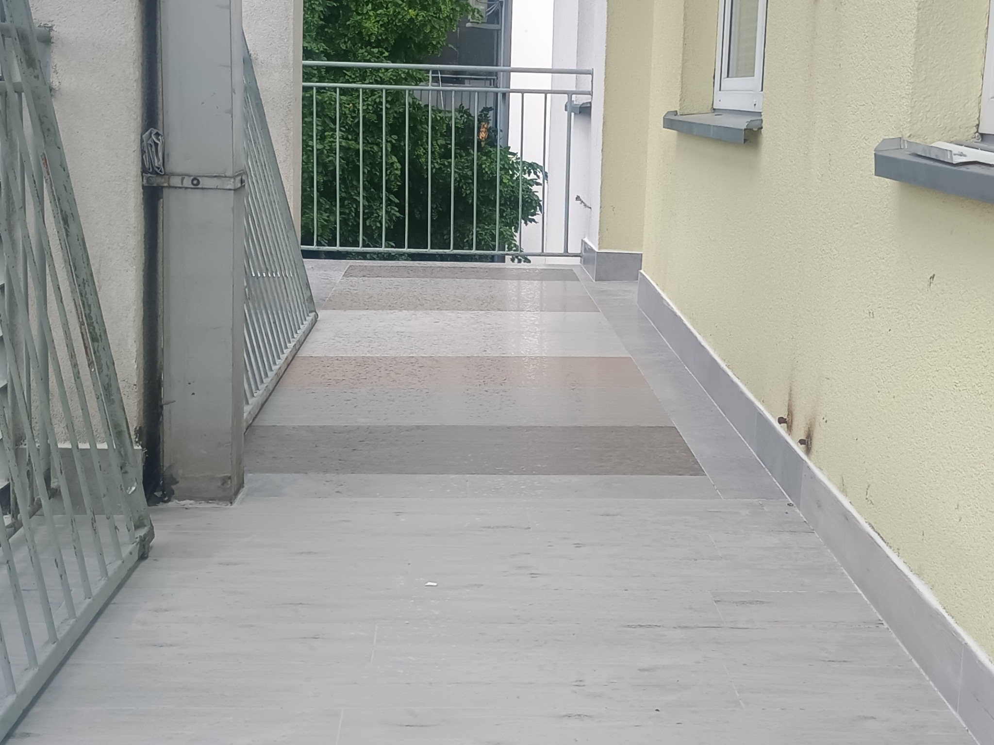 Balkon z nową, szarą, ułożoną płytką. Widoczne barierki i fragment elewacji budynku w jasnym odcieniu. Zastosowano różne odcienie szarości.